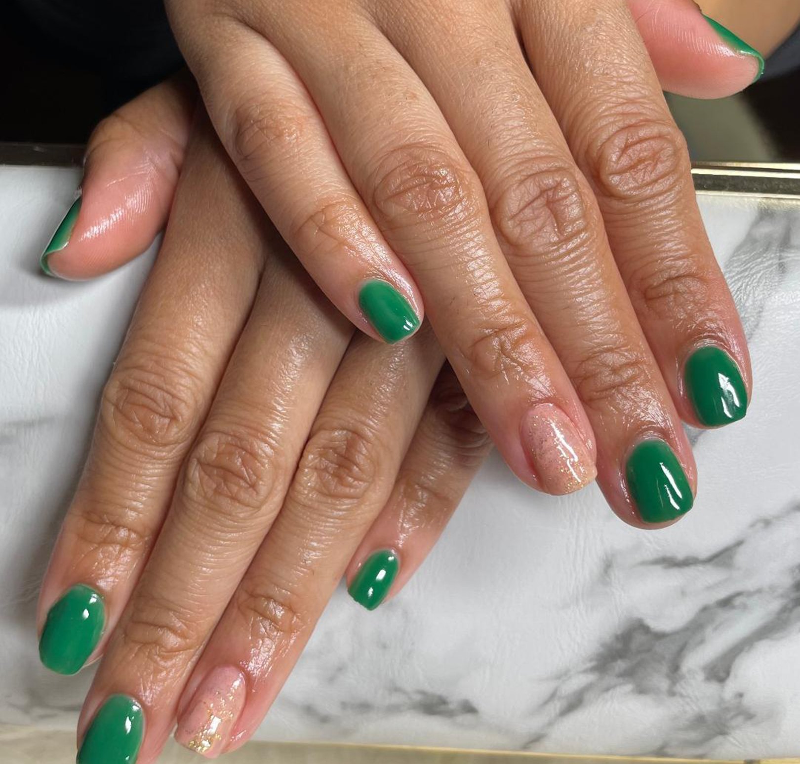 Hands of a laina woman on top of each other on a table with mylar on her ring finger nails and green color gel polish on the rest of the finger nails of borth hands